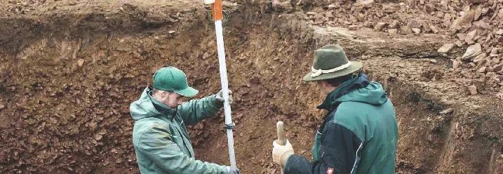 Zwei Personen vermessen Baugrube mit Messlatte.